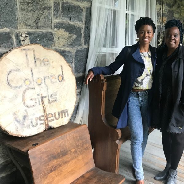 Two Black women stand next to each other to the right side of a wooden sign stating "The Colored Girls Museum"