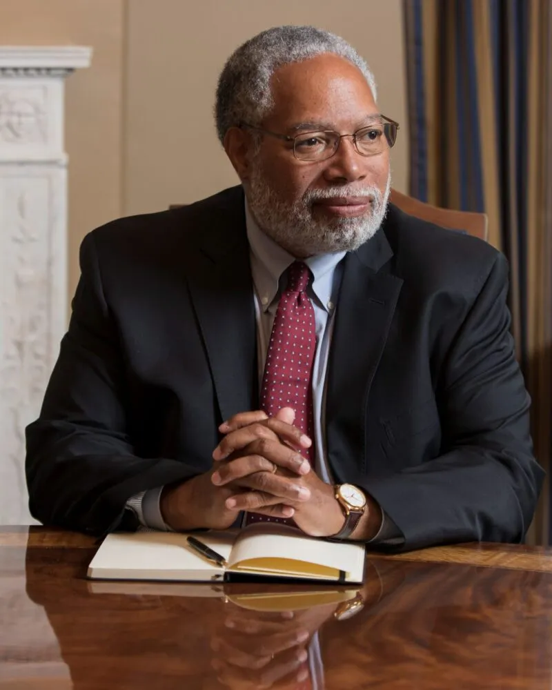 Portrait of Dr. Lonnie G. Bunch III, Secretary of the Smithsonian, a Black man with close cropped salt and pepper hair, glasses, and a mustache and beard, seated at a desk with an open notebook and pen, hands clasped on the desk, wearing a dark suit red tie, light colored shirt, a gold watch, with an architectural feature and curtain in the background