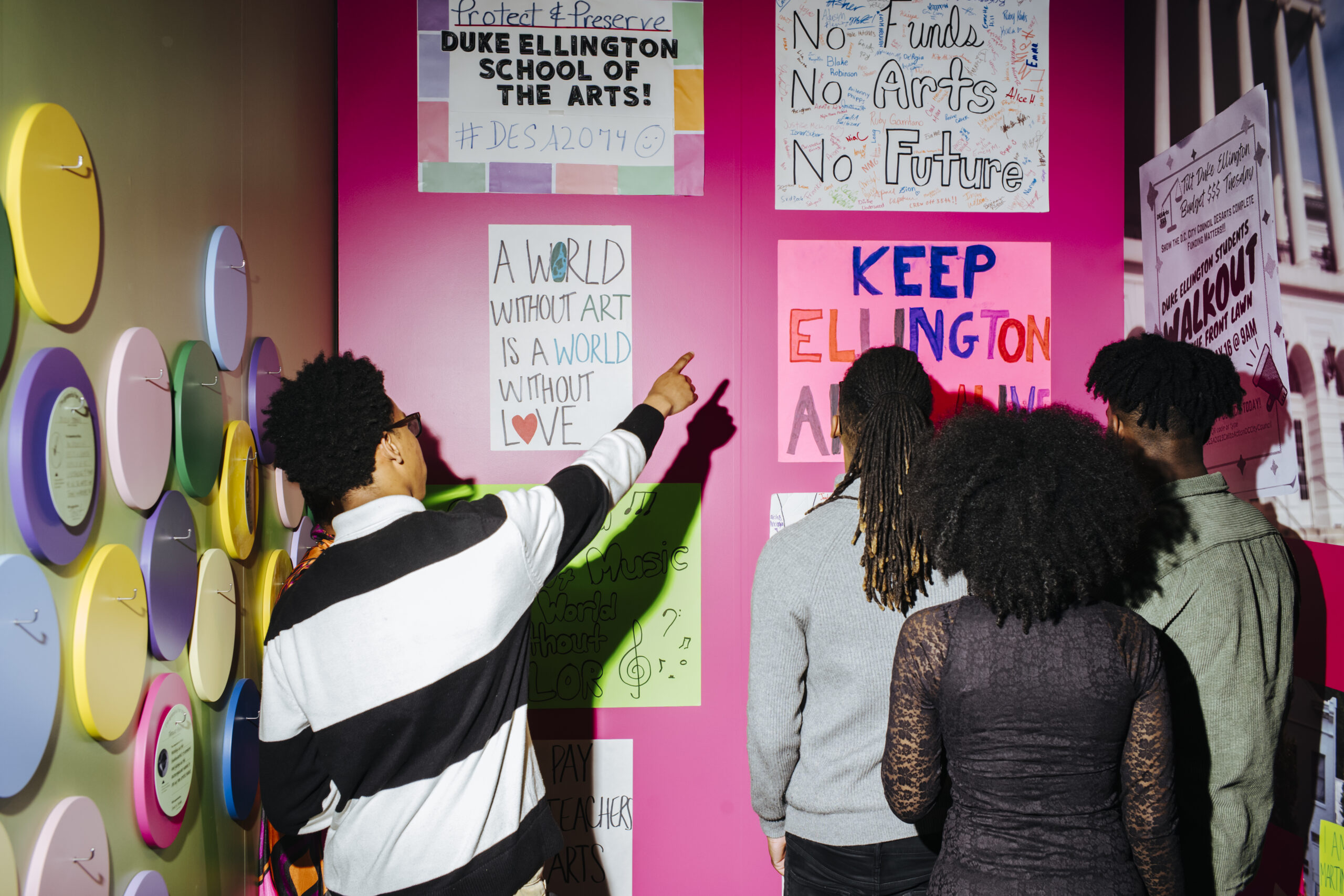 Three women of color with their backs to the viewer, one of whom points to a protest sign placed on a bright pink colored wall