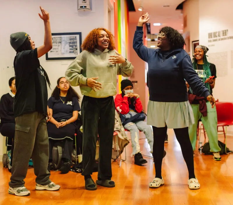 A group of women of color in a hallway, a few seated or standing in the back with three figures in the foreground standing and expressively gesturing to one another