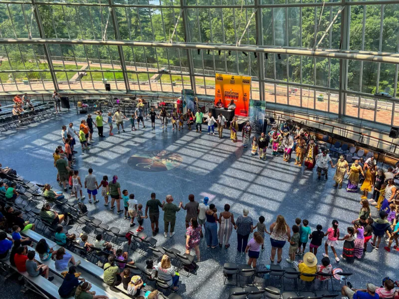 In an airy modern building with high ceilings and a wall of windows looking out onto trees, a crowd of all ages gathers in a large circle holding hands with additional onlookers seated in folding chairs at the sides, and a large poster with an orange and red gradient says 